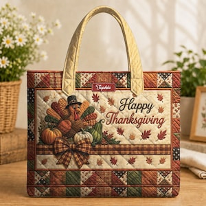 Op de afbeelding: Een gewatteerde draagtas met een Thanksgiving-thema, een kalkoen met een hoed, pompoenen en herfstbladeren. De tekst "Happy Thanksgiving" en de naam "Tophia" staan op de voorkant. De tas heeft gouden handvatten en een geruite rand.