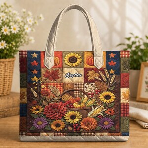 Op de afbeelding: Een gewatteerde tote bag met een patchworkontwerp met herfstthema, waaronder zonnebloemen, pompoenen, maïs en herfstbladeren. In het midden staat de naam Sophia in witte letters. De tas heeft twee lichtgekleurde, gewatteerde handvatten.