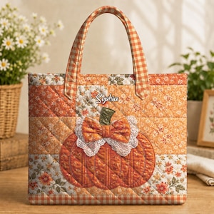 Op de afbeelding: Een gewatteerde tas met een patchworkontwerp in tinten oranje, wit en bruin. De voorkant toont een grote stoffen pompoen met een witte kanten rand en een strik, met de naam Sophia erboven geborduurd. De tas heeft twee handvatten met ruitpatroon.
