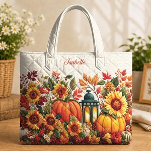 Op de afbeelding: Een witte gewatteerde tas met een levendige herfstprint van zonnebloemen, pompoenen, bessen en een lantaarn. De naam Isabelle is in schrijfletters bovenaan in het midden geborduurd. De tas heeft twee stevige hengsels en een ruitvormig stiksel.