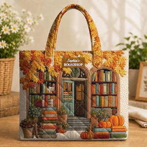 May include: A quilted tote bag featuring a printed scene of a bookshop entrance with shelves of books, autumn leaves, and pumpkins. The text "Sophia's BOOKSHOP" is printed on a rectangular sign above the door. The bag has two matching handles.