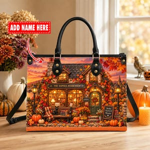 May include: A black handbag featuring a printed autumn scene of a bookstore named The Sophia Bookshop, with orange and red fall foliage, pumpkins, and a warm sunset sky. The bag has black handles and a rectangular shape with a customizable name area.