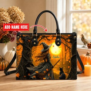 May include: A black handbag featuring a printed scene of a witch in a forest with a black cat and a large yellow moon. The bag has black handles and gold-tone hardware. The name Sophia is printed on the bag, and a red box says ADD NAME HERE.
