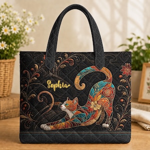 Op de afbeelding: Een zwarte gewatteerde tas met een illustratie van een uitrekkende kat en bloemmotieven in oranje, blauwgroen en rood. De naam Sophia is in goudkleurige letters aan de linkerkant geborduurd en de tas heeft twee handvatten.