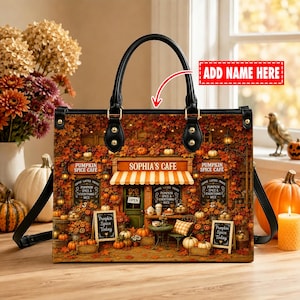 May include: A structured handbag featuring a printed autumn cafe scene with orange and brown foliage, a striped awning, and signs reading "Sophia's Cafe" and "Pumpkin Spice Cafe." The bag has black handles, gold-tone hardware, and a red "ADD NAME HERE" label.