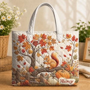 Könnte beinhalten: Eine gesteppte Tragetasche mit weißen Henkeln, die eine herbstliche Szene mit einem Eichhörnchen auf einem Ast, orangefarbenen und roten Ahornblättern, einem Holzzaun und zwei Kürbissen zeigt. Der Name Nancy steht unten rechts in roter Schreibschrift.