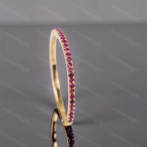 May include: A thin gold band ring featuring a continuous row of small, round, vibrant pink gemstones set along the outer edge. The ring is positioned vertically, showcasing the polished gold interior and the uniform setting of the pink stones.