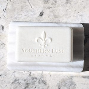 May include: A white bar of soap with the words "SOUTHERN LUXE - SAVON" and a fleur-de-lis design, resting on a white marble soap dish. The soap and dish are rectangular and have a clean, minimalist aesthetic.