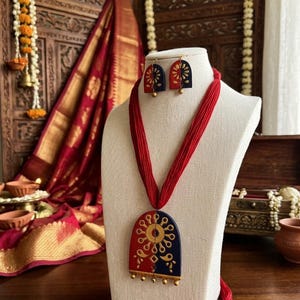 May include: A necklace and earring set featuring a semi-circular pendant and matching earrings in red and navy blue with gold-toned painted floral patterns. The necklace is suspended on a thick red thread cord and includes small gold-toned beads.