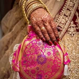 May include: A pink and gold floral embroidered pouch with a drawstring closure. The pouch is held by a hand decorated with henna and gold bangles. The nails are painted red. The background is a gold and maroon fabric.