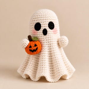 May include: A cream-coloured crocheted ghost figurine featuring two round black eyes, an oval black mouth, and soft pink cheeks. The ghost holds a small, bright orange crocheted pumpkin with a dark brown stem and a green leaf in its left arm.