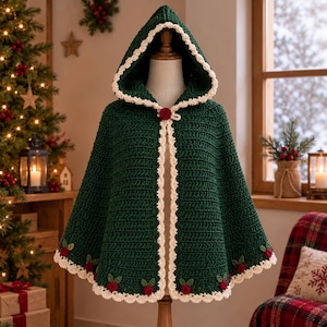 Könnte beinhalten: Ein dunkelgrüner Häkel-Umhang mit Kapuze und cremefarbenem, wellenförmigem Rand an den Kanten und der Kapuze. Das Kleidungsstück ist am unteren Saum mit kleinen gehäkelten roten Blumen- und grünen Blattmotiven sowie einer roten Blume am Hals verziert.