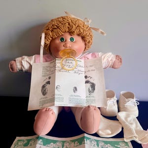 Può includere: Una bambola vintage Cabbage Patch Kids con capelli biondi di lana, occhi verdi e un ciuccio. La bambola indossa un completo rosa e bianco e tiene in mano un certificato di nascita. Accessori: scarpe e calzini bianchi.