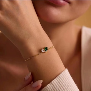 Puede incluir: Una delicada pulsera de cadena en tono dorado con dos piedras en forma de pera colocadas una al lado de la otra. Una piedra es transparente e incolora, mientras que la otra es de un vibrante color verde esmeralda. Diseño de eslabones finos.