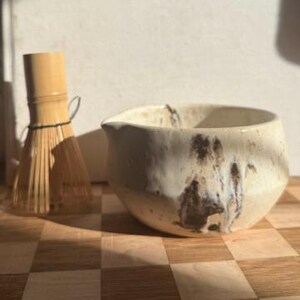 Könnte beinhalten: Eine Keramik-Matcha-Schale mit strukturierter, cremefarbener Glasur und braunen Akzenten, neben einem Bambusbesen. Die Schale hat eine unregelmäßige Form und einen kleinen Ausguss. Die Artikel stehen auf einer karierten Holzoberfläche.