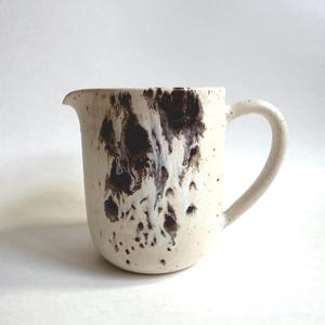May include: A ceramic pitcher with a cream-colored base featuring dark brown, ink-like splatters and drips concentrated on the front. The vessel has a rounded body, a small pouring spout, and a curved handle, with small brown speckles throughout.