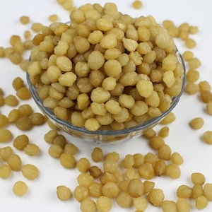 Puede incluir: Un cuenco de vidrio transparente lleno de pequeños alimentos redondos de color amarillo claro. Algunos de los alimentos están esparcidos alrededor del cuenco sobre una superficie blanca. Los alimentos parecen ser un aperitivo o un ingrediente.