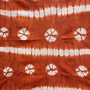 May include: Close-up of an orange fabric with white horizontal stripes and circular, star-like patterns. The fabric has a textured appearance, with visible stitching. The design is symmetrical, creating a visually appealing textile.