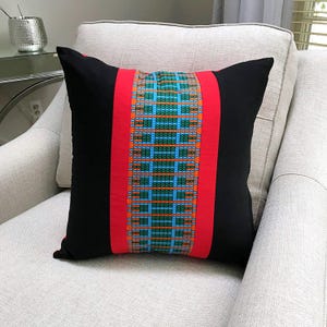 May include: A square throw pillow featuring a central vertical panel with a geometric pattern in blue, green, and orange. This panel is flanked by two solid red vertical stripes, with the remaining side sections of the pillow cover in solid black.