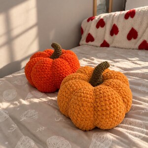 May include: Two round, crocheted pumpkin-shaped cushions. One pumpkin is a vibrant orange, while the other is a lighter, golden-orange shade. Both feature a textured, yarn-based surface and a short, dark brown crocheted stem at the centre top.