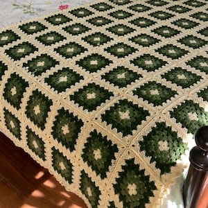 May include: A crochet blanket featuring a repeating granny square pattern in shades of forest green, sage green, and cream. The square motifs are joined together to form a large textile piece with a textured, open-work yarn construction.