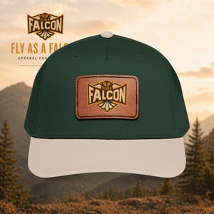 Può includere: Cappellino da baseball verde scuro con visiera beige e toppa in pelle. La toppa presenta la scritta "FALCON" in lettere dorate, con la scritta "FLY AS A FAL" sopra. Il cappellino è abbigliamento per gli amanti dell'outdoor.