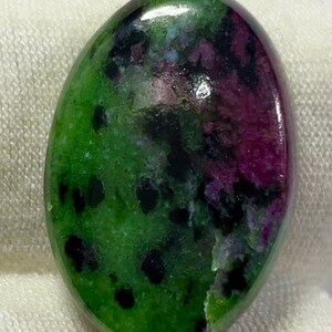 Può includere: Una pietra preziosa lucidata di forma ovale caratterizzata da una base di colore verde vibrante con macchie distinte di magenta intenso e inclusioni nere sparse su tutta la superficie, che presenta una finitura liscia, riflettente e lucida.