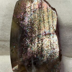 May include: A polished sunstone gemstone featuring a translucent brown base with a vibrant, iridescent aventurescence effect. The surface displays a shimmering, multi-coloured metallic lustre with flecks of gold, green, and pink light reflections.