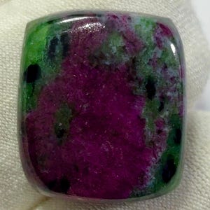 Peut inclure: Une pierre précieuse cabochon polie de forme coussin, présentant un centre magenta-rose vibrant entouré d'inclusions vert lime et noir foncé. La surface est lisse et réfléchissante, mettant en valeur un motif minéral naturel multicolore.