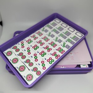 May include: A Mahjong set with white tiles featuring red, green, and black symbols, arranged in a purple tray. The tray is stacked on another purple tray, with pink paper and additional tiles visible.