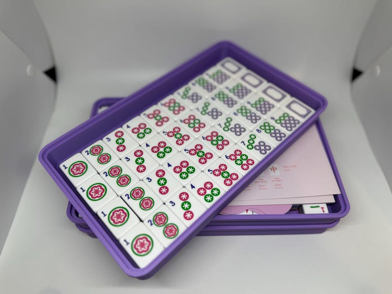 May include: A Mahjong set with white tiles featuring red, green, and black symbols, arranged in a purple tray. The tray is stacked on another purple tray, with pink paper and additional tiles visible.