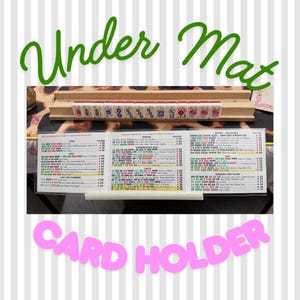 May include: A wooden Mahjong tile rack holding a row of fourteen tiles with various symbols and colors. Below the rack sits a tri-fold Mahjong score card with printed text and numerical values. The text Under Mat and CARD HOLDER is visible.