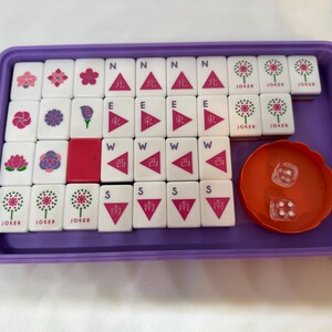 May include: A Mahjong set in a purple tray. The set includes white tiles with various symbols and characters, along with an orange dice cup and two dice. The tiles feature floral designs, directional symbols, and the word "JOKER".