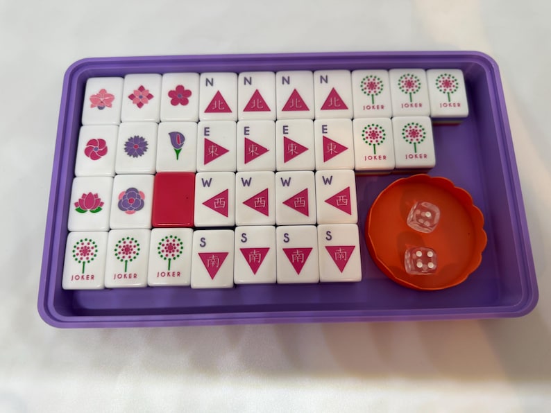 May include: A Mahjong set in a purple tray. The set includes white tiles with various symbols and characters, along with an orange dice cup and two dice. The tiles feature floral designs, directional symbols, and the word "JOKER".