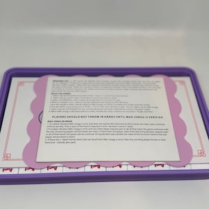 May include: A purple rectangular tray contains a Mah Jongg set. The set includes tiles and a rules sheet with text. The rules sheet is light pink with a scalloped edge. The tiles are white with purple markings.