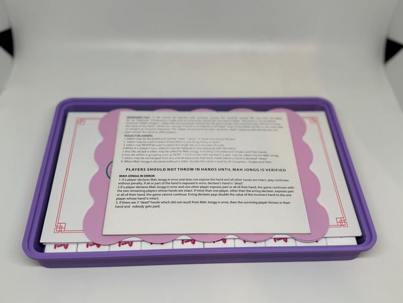 May include: A purple rectangular tray contains a Mah Jongg set. The set includes tiles and a rules sheet with text. The rules sheet is light pink with a scalloped edge. The tiles are white with purple markings.