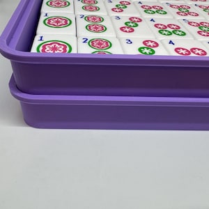 May include: A purple plastic tray containing white Mahjong tiles. The tiles feature red and green floral patterns and black numbers. The rectangular tray has a raised edge. The tiles are arranged in an orderly fashion within the tray.
