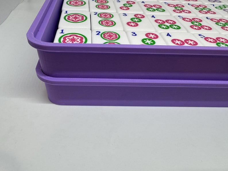 May include: A purple plastic tray containing white Mahjong tiles. The tiles feature red and green floral patterns and black numbers. The rectangular tray has a raised edge. The tiles are arranged in an orderly fashion within the tray.