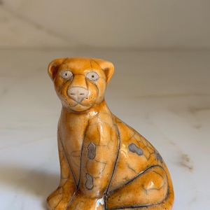 May include: A ceramic figurine of a seated cheetah with an orange and tan glaze. The cheetah has a detailed face with dark eyes and a light-colored nose. The figurine has a cracked glaze effect and dark lines that outline the cheetah's form.