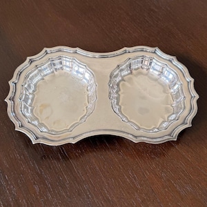 May include: A silver-toned metal double dish tray featuring two circular, scalloped-edge bowls connected by a central bridge. The entire piece has a reflective, polished finish with a decorative, wavy, and pointed outer rim border design.