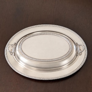 May include: An oval silver-toned serving platter with a raised central section and two curved handles on the sides. The rim and the inner border of the platter feature a detailed, repeating floral or vine-like embossed pattern in relief.