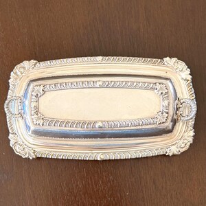 May include: A rectangular silver-toned serving tray featuring an ornate, raised border with intricate leaf and scroll detailing. The tray includes two small, decorative handles on the shorter ends and a smooth, recessed central rectangular panel.