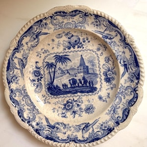 May include: A round ceramic plate featuring a blue and white transferware pattern. The center depicts a landscape with palm trees, a building, and figures with camels. The border is decorated with floral motifs and birds on a cream background.