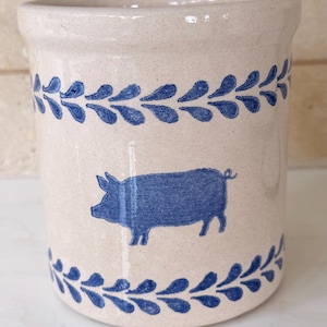 May include: A cylindrical ceramic crock with a speckled off-white finish. The exterior features a hand-painted blue pig silhouette centered between two horizontal bands of blue leaf-like patterns encircling the top and bottom edges.