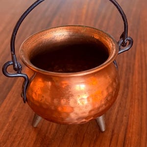 May include: A small, round, hammered copper cauldron featuring a dark metal swing handle attached to two side loops. The vessel is supported by three short, sturdy legs and has a wide, open top with a smooth, polished rim and a deep interior.