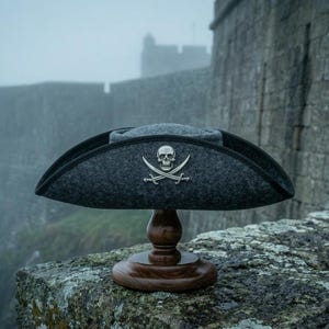 May include: A charcoal gray felt tricorn hat featuring a silver-toned metal skull and crossbones emblem on the front. The hat is displayed on a polished dark brown wooden stand, showcasing its wide, curved brim and structured crown design.
