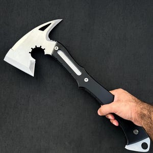 Puede incluir: Un hacha t&aacute;ctica con cabeza de metal plateado, hoja curva, punta afilada y un recorte circular con dientes tipo engranaje. El mango es negro mate con una incrustaci&oacute;n plateada, asegurado por dos tornillos, e incluye un orificio para cord&oacute;n.
