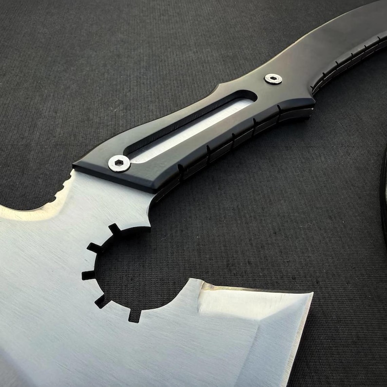 Puede incluir: Un hacha t&aacute;ctica con una hoja de metal plateado que presenta un recorte circular con dientes tipo engranaje. El mango est&aacute; construido con material negro con una incrustaci&oacute;n plateada central, asegurado por dos tornillos hexagonales visibles.