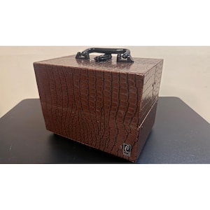 May include: A rectangular brown storage case with a textured crocodile skin pattern. The case features a black plastic handle on the top and a small silver-toned square emblem with a stylized letter C on the lower right corner of the front panel.