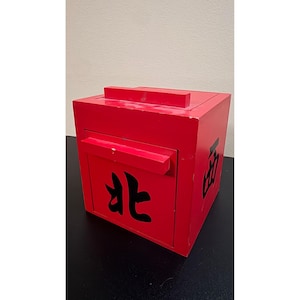Puede incluir: Una caja cuadrada de madera roja con tapa abatible y un cajón frontal. El panel frontal presenta el carácter chino negro para Norte, mientras que el panel lateral muestra el carácter chino negro para Oeste, ambos pintados en un estilo negrita.
